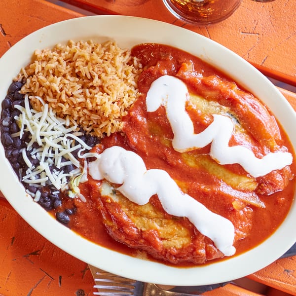 A Mexican dish with two enchiladas covered in red sauce and sour cream, rice, black beans, and shredded cheese.