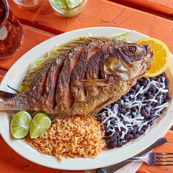 Whole fried fish with rice, black beans, cheese, lime, and orange on a white plate.