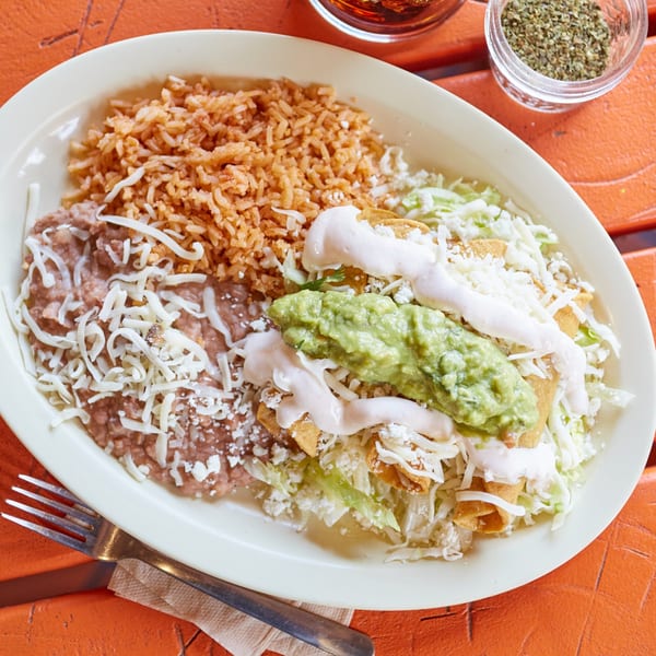 Mexican plate with rice, refried beans, taquitos, guacamole, sour cream, and shredded cheese.